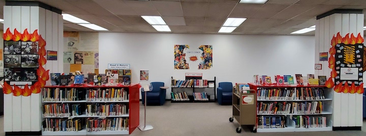 Photo of the SDSU Comics Corner showing a display of banned comics. The corner has two red bookshelves with an opening in the middle. The banned comics are displayed on top of the shelves. On either side of the shelves are columns with posters. There are flame (fire) decorations, information about book banning, and historic photos of children reading comic books with anti-censorship quotes. 